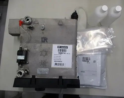 Wärmetauscher von Remeha, mit verschiedenen Anschlüssen und Zubehörteilen, auf einem Tisch platziert. Mehrere Plastiktüten und Flaschen befinden sich im Bild.